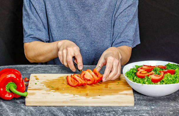 Vrouwelijke handen snijden tomaat op snijplank. Blauw t-shirt, zwarte achtergrond. Saladebereiding - Foto, afbeelding