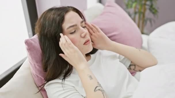 Uitgespannen jonge vrouw, in pyjama, zittend op slaapkamer bed met de hand op het hoofd, pijn lijdend aan migraine - Video