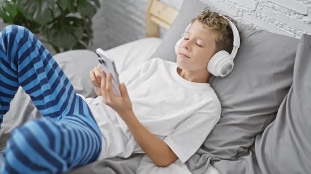 Roztomilý blonďatý chlapec šťastně surfuje na internetu, sedí bdělý na posteli v ložnici, odpočívá v pyžamu, sebevědomě sleduje video na smartphonu, domácí atmosféra ho rozesměje. - Záběry, video