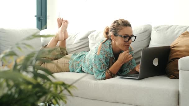  ソファーの上に横たわる現代の女性は,リラックスしてノートパソコンを使ってウェブや仕事を閲覧します. 屋内レジャーアクティビティで時間と技術の接続を楽しんでいる人.  - 映像、動画