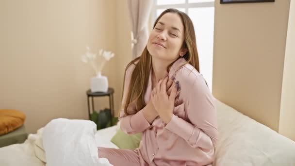 Vrolijke jonge vrouw met blauwe ogen, zittend op haar bed in pyjama, dankbaar lachend en hand in hand op de borst in een gezonde ochtend gebaar in haar kamer - Video