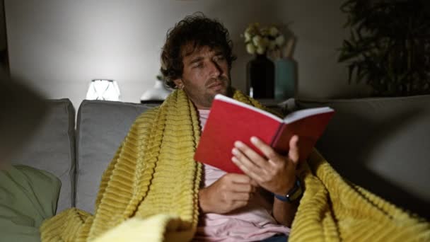 Schöner Mann, der es sich zu Hause mit einem Buch auf dem Sofa gemütlich macht und Ruhe in einem gemütlichen Innenraum ausstrahlt. - Filmmaterial, Video