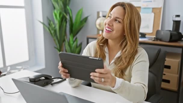 Hymyilevä latino nainen nauttii työskentelystä tabletin kanssa modernissa toimistossa. - Materiaali, video