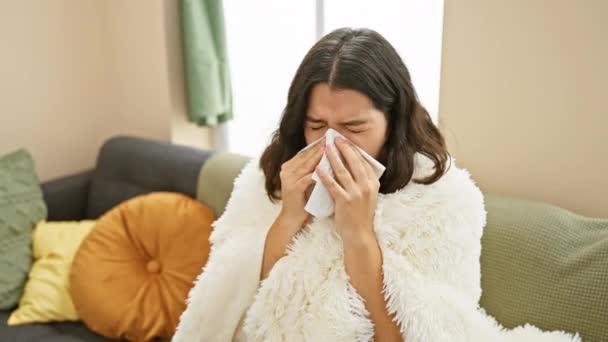 Eine junge hispanische Frau pustet sich mit einem Taschentuch in die Nase, während sie zu Hause auf einem Sofa sitzt und unwohl aussieht. - Filmmaterial, Video