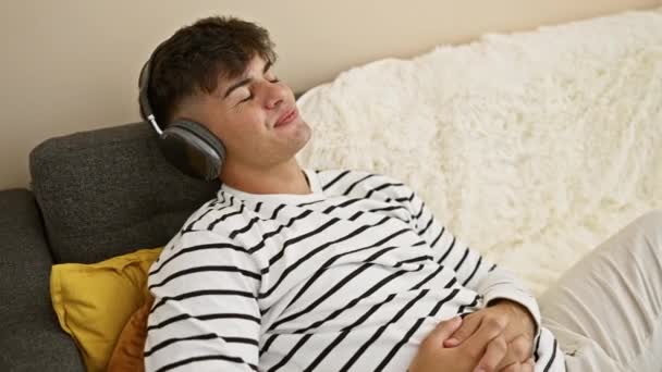 Alegre joven hispano en zona de auriculares escuchando música bombeada, disfrutando con confianza del divertido estilo de vida interior. él está reclinado en un sofá en casa, lleno de felicidad y sonriendo. - Imágenes, Vídeo