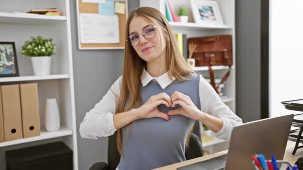 Joven mujer alegre haciendo signo de forma de corazón con los dedos en la oficina, trabajador de negocios rubia irradia felicidad, sonriendo en el interior, simbolizando el amor y la amistad - Metraje, vídeo