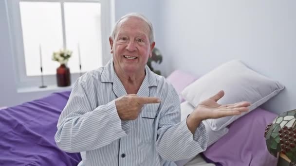 Vrolijke oudere man, verbaasd terwijl hij in pyjama op bed zat, vol vertrouwen met de hand presenterend, wijzend en glimlachend in de huiselijke sfeer van zijn slaapkamer. - Video