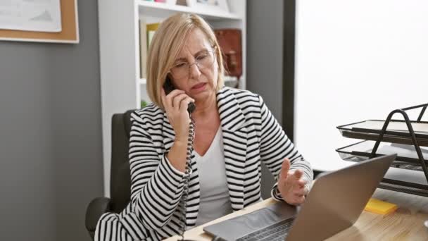 Aggódó érett nő beszél telefonon az irodában íróasztal laptop és dokumentumok - Felvétel, videó