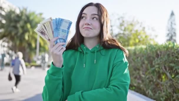 Young woman holding hungarian forint in a sunny park, looking thoughtful and smiling in urban surroundings. - Footage, Video