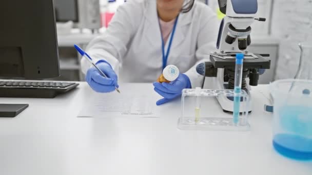 Сфокусована іспаномовна жінка вчений вивчає пляшку ліків в лабораторії з мікроскопом і пробірками поблизу. - Кадри, відео