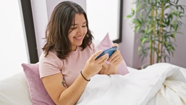 Mujer hispana riendo mientras juega en smartphone en dormitorio luminoso - Imágenes, Vídeo