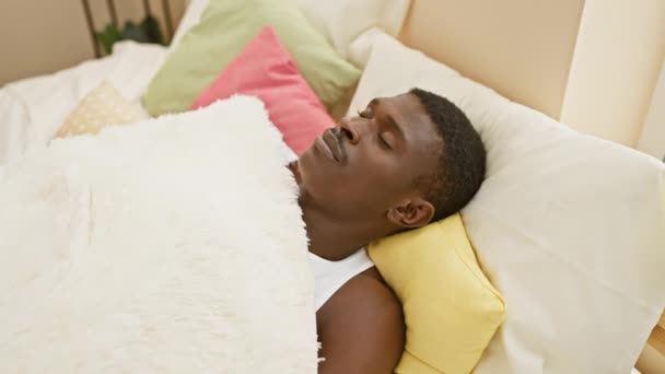 Afrikalı bir adam rahat bir yatak odasında, huzurlu bir ifadeyle renkli yastıkların üzerinde uzanıyor. - Video, Çekim