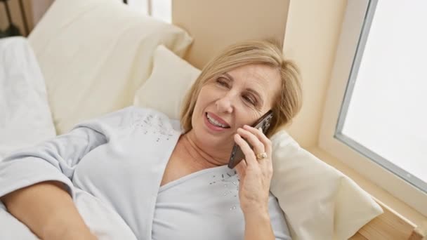 Een glimlachende vrouw van middelbare leeftijd praat aan de telefoon terwijl ze zich ontspant in haar slaapkamer, en laat een comfortabel, casual lifestyle moment zien. - Video