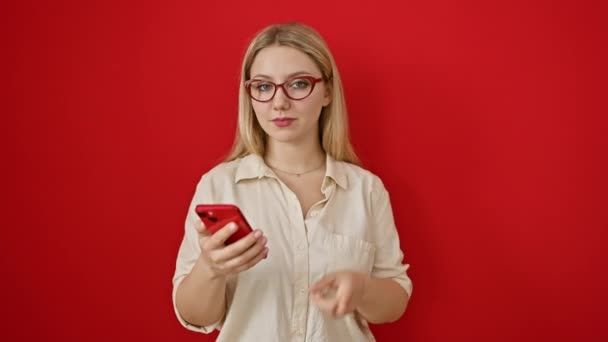 Vidám szőke fiatal nő vidáman mutatva és mosolyogva az egyik oldalon a piros háttér SMS-ezés közben, portré vonzó hölgy telefonnal a kezében - Felvétel, videó