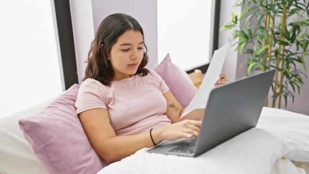 Una mujer hispana enfocada trabaja en su computadora portátil en un acogedor dormitorio, papeles en la mano, equilibrando la productividad y la comodidad. - Imágenes, Vídeo