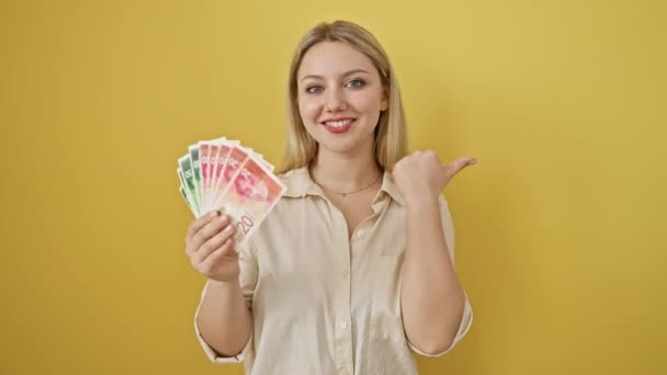 Joyful jonge vrouw knippert een duim omhoog, grijnzend met bankbiljetten van Israël shekel in de hand, op geïsoleerde gele muur - Video