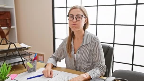 Hilarious office caper, young business woman sitting, amusingly puffing cheeks, inflating mouth with air, making funny face. breath held, eyes crazy in the indoor workplace. - Footage, Video