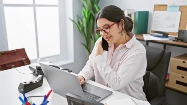 Genç bir İspanyol iş kadını telefonda gülümseyen ve dizüstü bilgisayar kullanan bir ofis ortamında birden fazla iş yapıyor.. - Video, Çekim