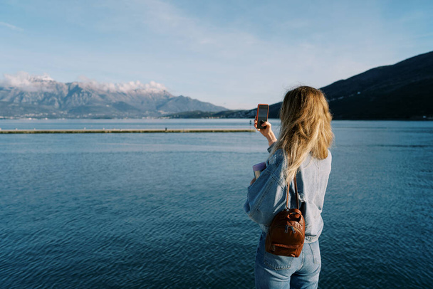 Kadın kıyıda dururken bir deniz ve dağ sırasını akıllı telefonuyla vuruyor. Arkadan bak. Yüksek kalite fotoğraf - Fotoğraf, Görsel