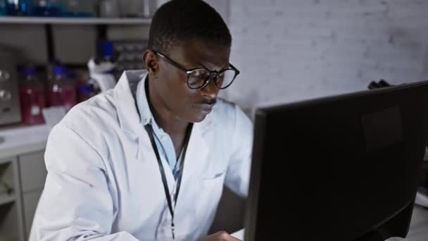 Ένας εστιασμένος Αφρικανός άνδρας σε ένα εργαστήριο φορώντας γυαλιά και ένα λευκό παλτό δουλεύει εντατικά σε έναν υπολογιστή.. - Πλάνα, βίντεο