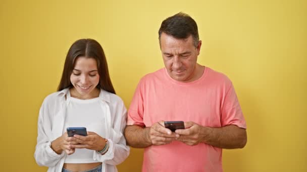 Ευτυχισμένος ισπανόφωνος πατέρας και κόρη μοιράζονται τη λάμψη των smartphones τους, στέλνοντας χαρούμενα μηνύματα και κρατώντας μια ψηφιακή συνομιλία σε ένα φωτεινό, απομονωμένο κίτρινο φόντο - Πλάνα, βίντεο