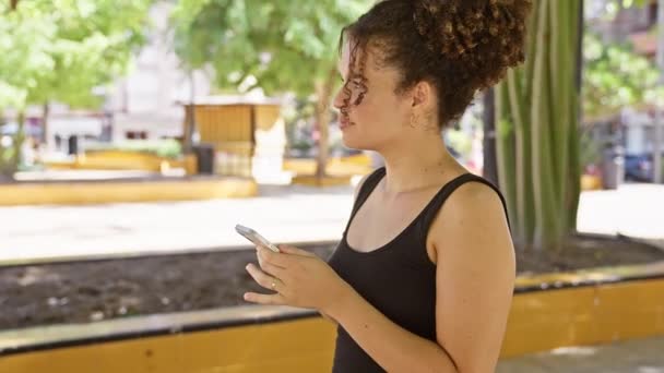 Μια νεαρή γυναίκα με σγουρά μαλλιά χρησιμοποιεί το smartphone της ενώ στέκεται σε ένα καταπράσινο πάρκο. - Πλάνα, βίντεο