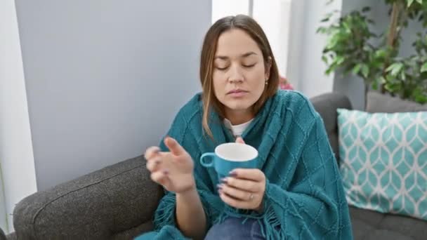 Een vrouw gewikkeld in een blauwe deken nipt thee op een bank binnen, die rust en comfort uitdrukt. - Video