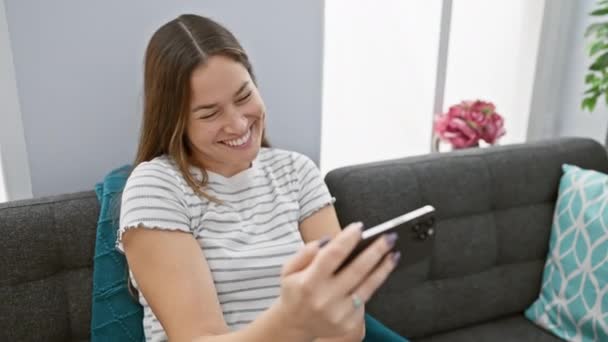 Joyeux jeune femme avec de longs cheveux bruns en utilisant un smartphone sur un canapé à la maison, exprimant joie et rire. - Séquence, vidéo