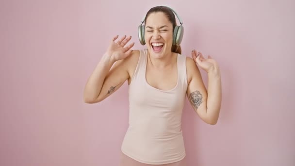 スポーツウェアの青い目を持つエキサイティングな若いブルネットの女性は,ヘッドフォンで音楽を聴く喜びの勝利を祝い,ピンクの孤立した背景の上に勝利を叫びます! - 映像、動画