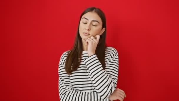 Jonge mooie Latijns-Amerikaanse vrouw dragen strepen t shirt staande denken geconcentreerd over twijfel met de vinger op de kin en kijken omhoog afvragen over geïsoleerde rode achtergrond - Video