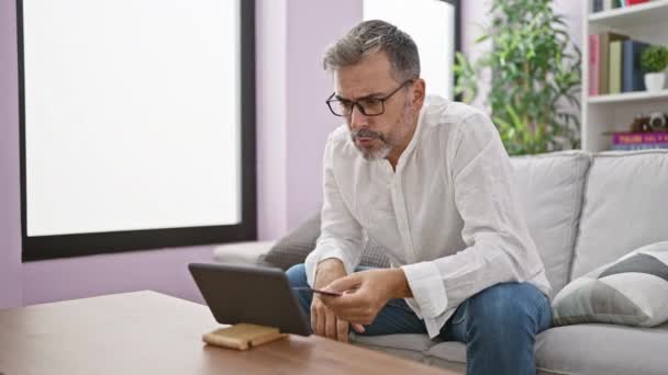 Jeune homme hispanique aux cheveux gris attrayant, impliqué dans une vidéoconférence financière sérieuse, assis à la maison sur le canapé, tenant une carte de crédit, plongé dans une conversation en ligne sur l'investissement des entreprises. - Séquence, vidéo