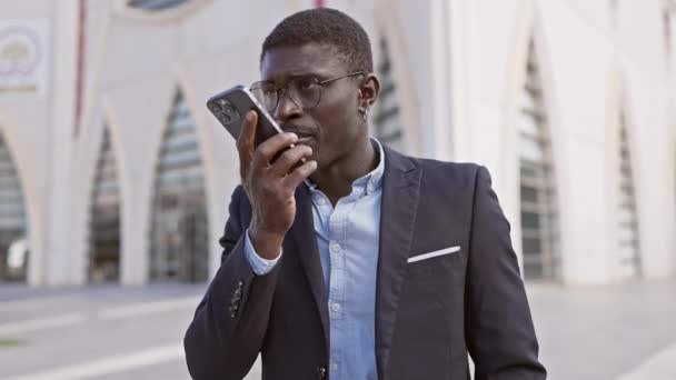Een Afrikaans-Amerikaanse man in professionele kleding maakt gebruik van een smartphone op een zonnige straat. - Video