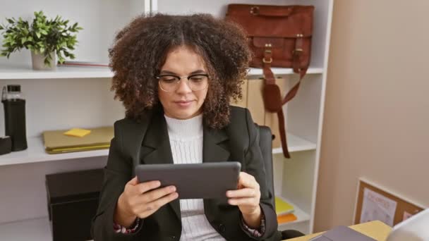 Une jeune femme hispanique aux cheveux bouclés portant des lunettes et des vêtements de travail travaille sur une tablette dans un environnement de bureau moderne. - Séquence, vidéo