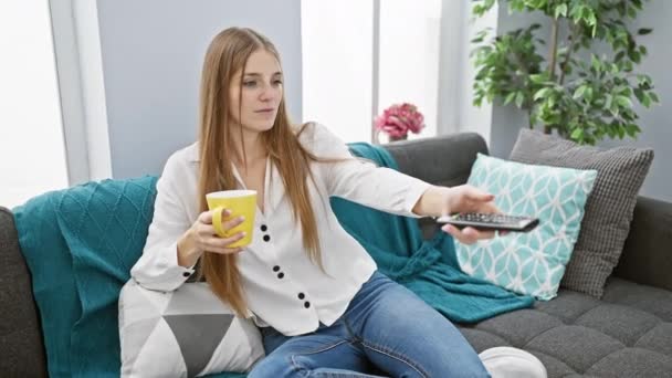 Una joven mujer caucásica se relaja con una bebida en un sofá, viendo la televisión en un acogedor salón. - Metraje, vídeo
