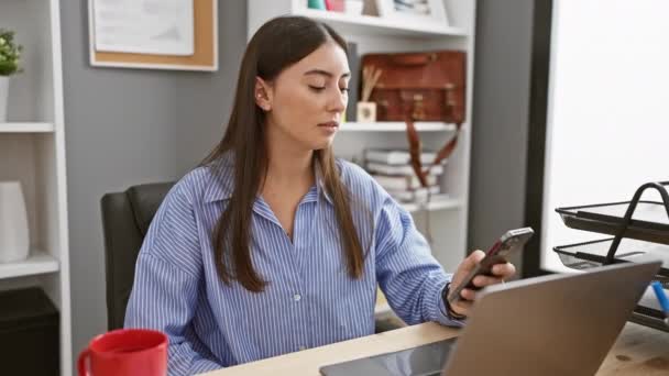 Latinalaisamerikkalainen nainen käyttää älypuhelinta työskennellessään kannettavan tietokoneen kanssa nykyaikaisessa toimistoympäristössä. - Materiaali, video