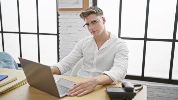 Vrolijk en zelfverzekerd, jonge Kaukasische man die op zijn laptop werkt in een kantoorruimte. zakenman spijkert zijn baan met onwankelbaar vertrouwen en een stralende glimlach. - Video