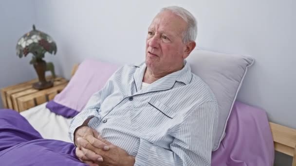 Oudere man in pyjama fronst sceptisch op bed, toont negatieve emotie als gevolg van een probleem. zenuwkarakter portrettering in slaapkamer setting. - Video