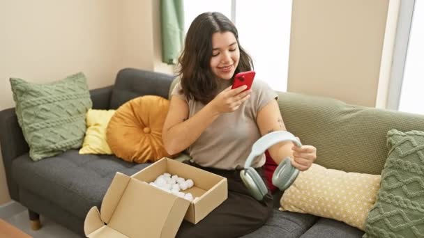 Spanyol nő mosolyog telefonnal, unbox fülhallgató kényelmes nappaliban, kifejező öröm - Felvétel, videó