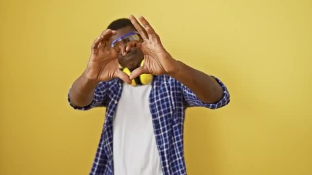 Jóképű afro-amerikai férfi biztonsági szemüvegben áll szemben egy elszigetelt sárga háttérrel, jelképes szív alakú kézmozdulattal mutatja a szerelmet, boldogságot sugároz ebben a romantikus koncepcióban.. - Felvétel, videó