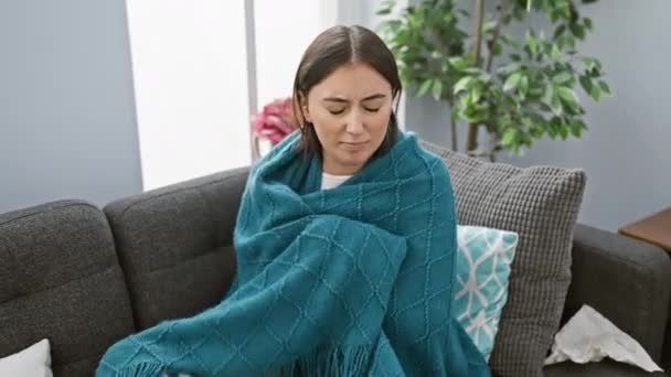 Hispanic woman feeling unwell, wrapped in turquoise blanket on sofa indoors, looking tired and sick. - Footage, Video