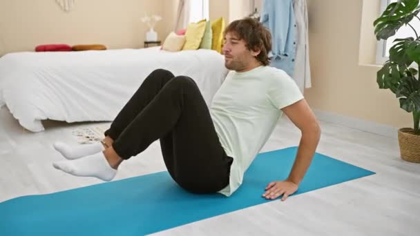 Sıradan giyinmiş genç sakallı bir adam yatak odasındaki mavi bir paspasın üzerinde karın egzersizi yapıyor. Bu da ev içi egzersizlerin sağlıklı olduğunu gösteriyor.. - Video, Çekim