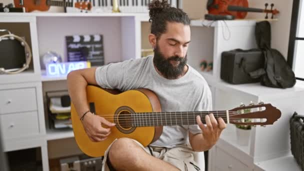 Hombre hispano con barba tocando la guitarra en el interior, sonriendo en un estudio de música. - Metraje, vídeo