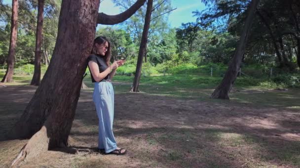 Bir kadın büyük bir çam ağacının gölgesinde cep telefonu kullanarak rahatça ayakta duruyor. - Video, Çekim