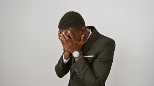 Uomo afroamericano depresso in giacca e cravatta che nasconde la sua triste espressione con una faccia piangente, coprendola con le mani su uno sfondo bianco isolato - Filmati, video