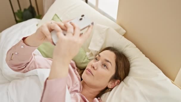 Μια casual, γαλήνια γυναίκα ξαπλωμένη στο κρεβάτι, χρησιμοποιώντας το smartphone της σε ένα φωτεινό, άνετο περιβάλλον υπνοδωμάτιο. - Πλάνα, βίντεο