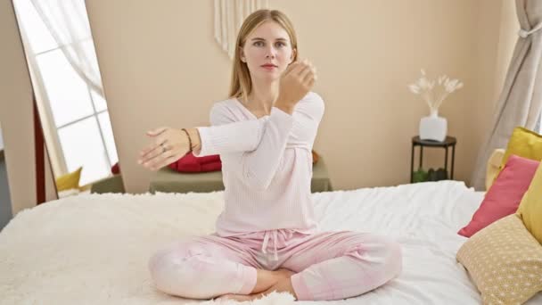 Mujer rubia en pijama rosa practicando yoga en una cama en un ambiente acogedor dormitorio. - Metraje, vídeo