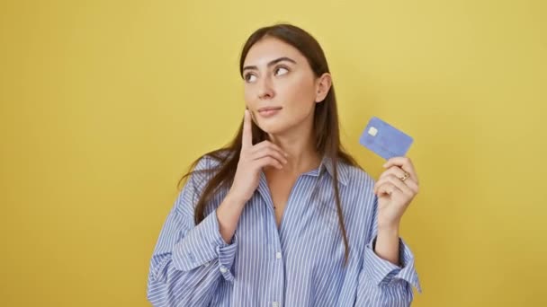 Young and beautiful hispanic woman surprised with a brilliant idea, joyfully points finger holding credit card, isolated on yellow background - Footage, Video