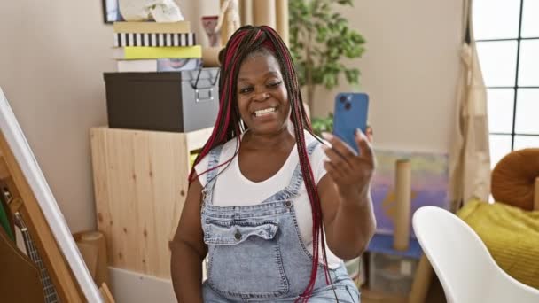 アフリカ系アメリカ人女性アーティスト,彼女のアートスタジオに座って,彼女のスマートフォンでビデオ通話をすることによって美しくテクノロジーを受け入れる. - 映像、動画