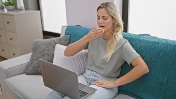 Une femme blonde fatiguée bâillant tout en travaillant sur son ordinateur portable assis sur un canapé gris à la maison, exprimant la fatigue. - Séquence, vidéo