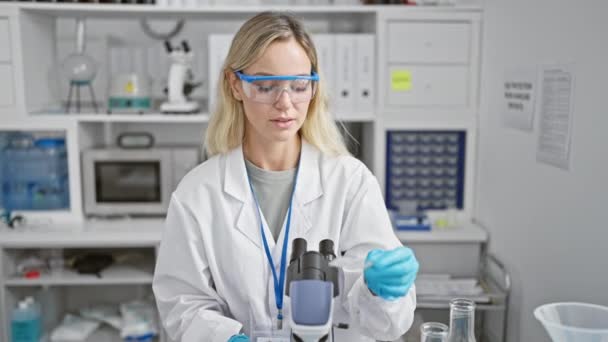 Une femme scientifique blonde concentrée utilisant un microscope dans un cadre de laboratoire moderne. - Séquence, vidéo